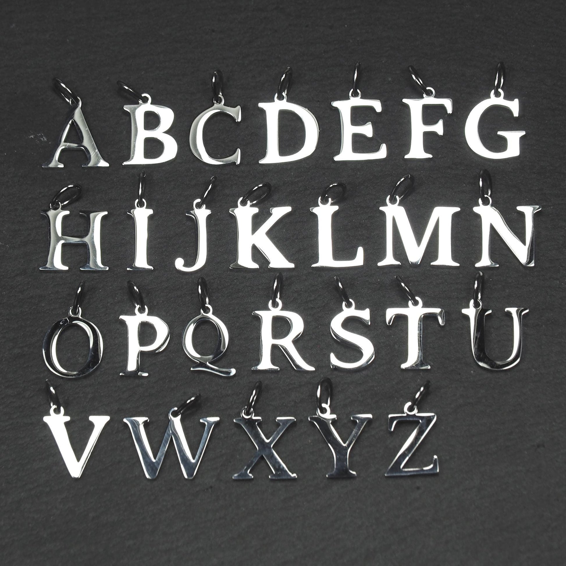 Buchstaben-Anhänger A–Z aus poliertem silberfarbenem Metall in serifenförmiger Typografie, mit Öse und Ring auf dunklem Grund.