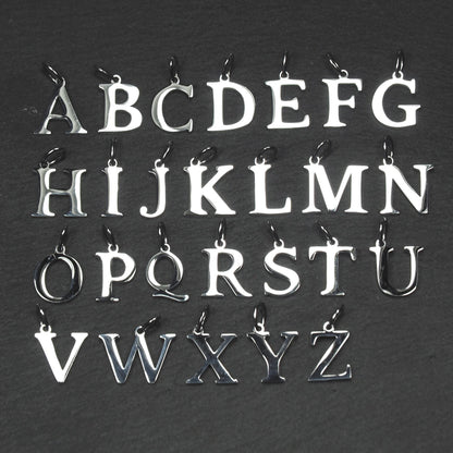 Buchstaben-Anhänger A–Z aus poliertem silberfarbenem Metall in serifenförmiger Typografie, mit Öse und Ring auf dunklem Grund.