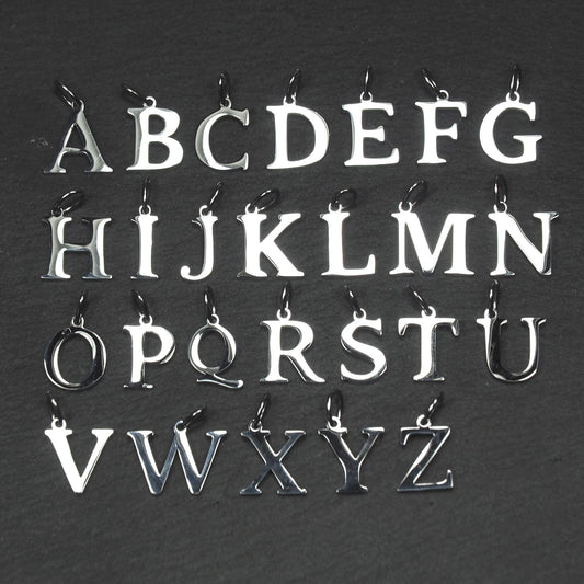 Buchstaben-Anhänger A–Z aus poliertem silberfarbenem Metall in serifenförmiger Typografie, mit Öse und Ring auf dunklem Grund.