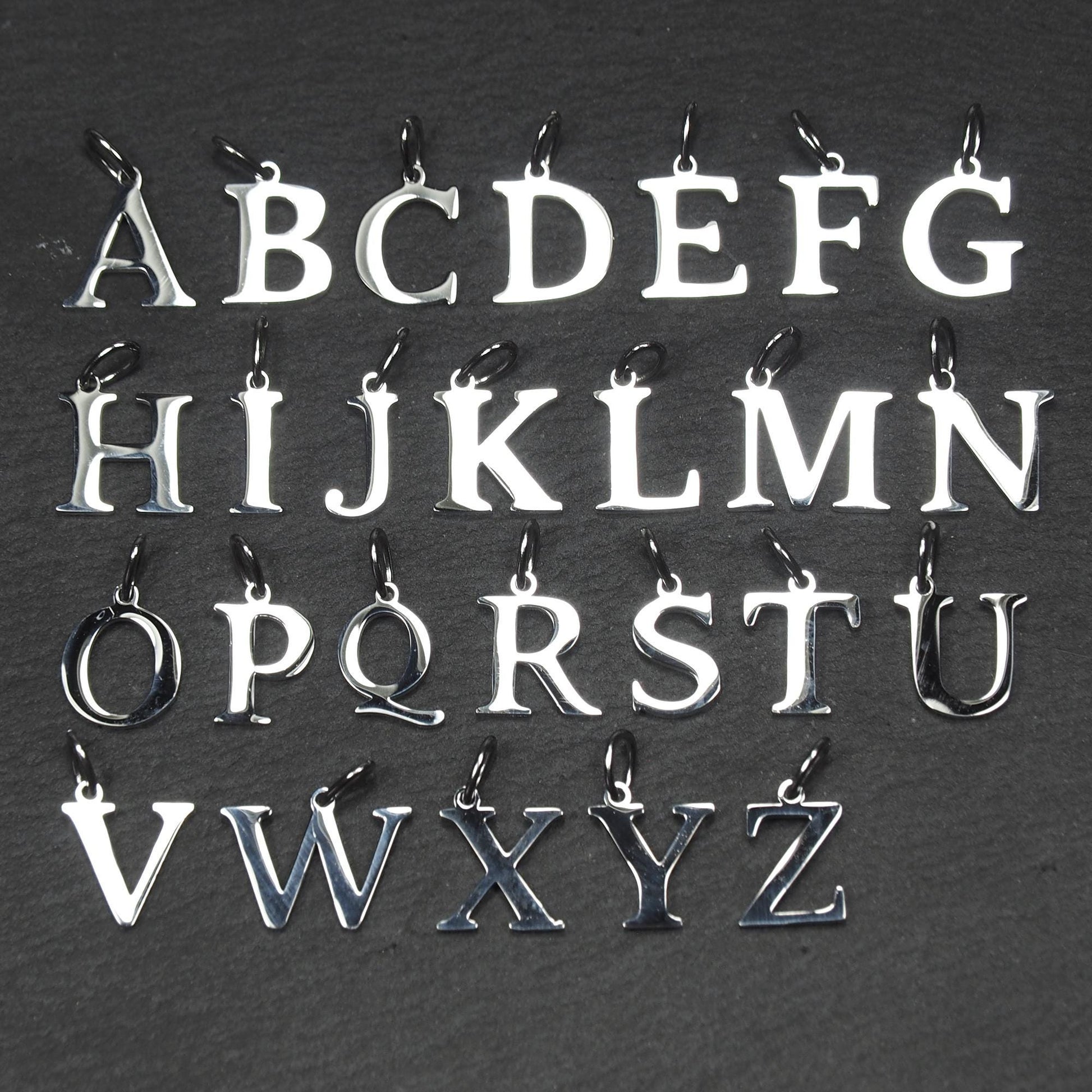 Buchstaben-Anhänger A–Z aus poliertem silberfarbenem Metall in serifenförmiger Typografie, mit Öse und Ring auf dunklem Grund.