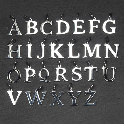 Buchstaben-Anhänger A–Z aus poliertem silberfarbenem Metall in serifenförmiger Typografie, mit Öse und Ring auf dunklem Grund.