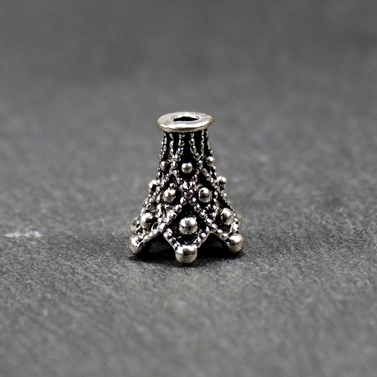 Konische, antik-silberne Perlenkappe mit durchbrochenem filigranem Ornament, reliefartigen Kugeln und sichtbarem Fädelloch oben.