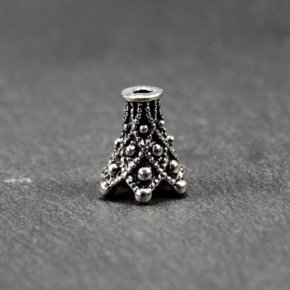 Konische, antik-silberne Perlenkappe mit durchbrochenem filigranem Ornament, reliefartigen Kugeln und sichtbarem Fädelloch oben.