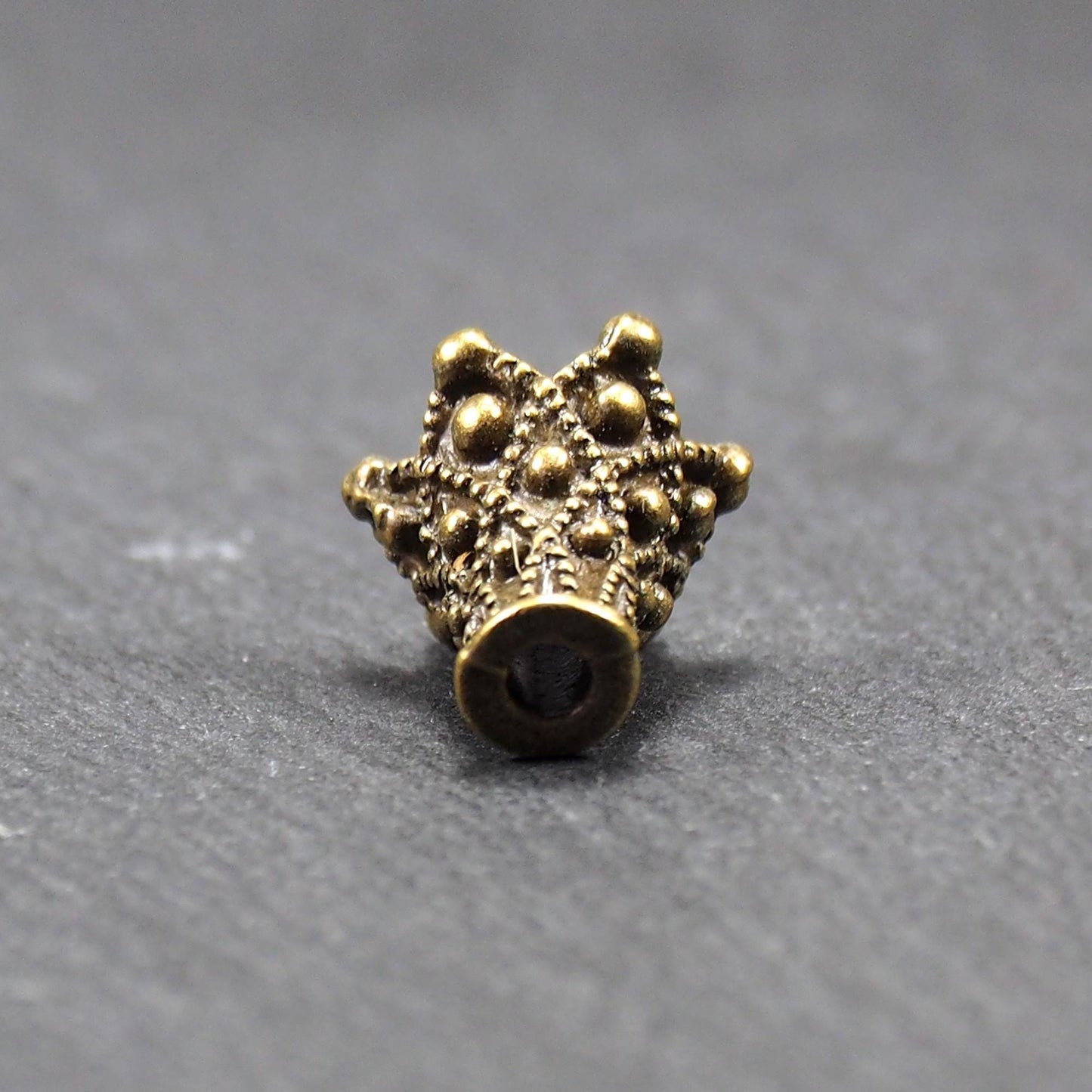 Konische bronzefarbene Perlenkappe aus Metall mit filigranem Ornamentreliéf, kugeligen Verzierungen und zentralem Fädelloch.