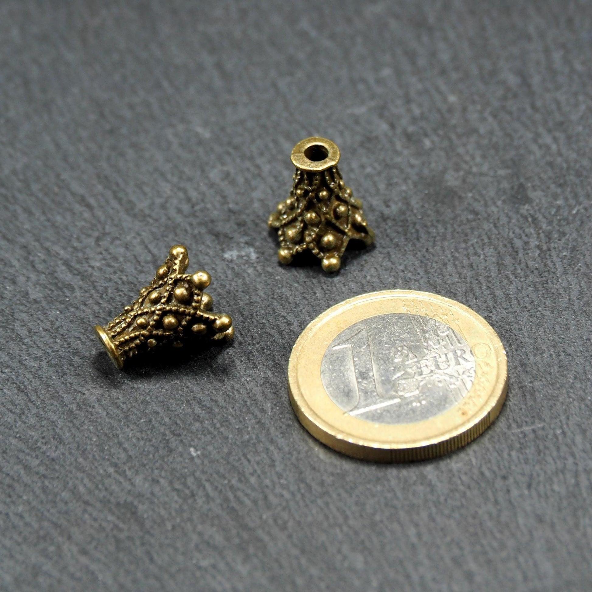 Zwei konische, antik-bronzefarbene Perlenkappen mit filigranem Relief und kleinen Kugelornamenten, neben einer 1-Euro-Münze.