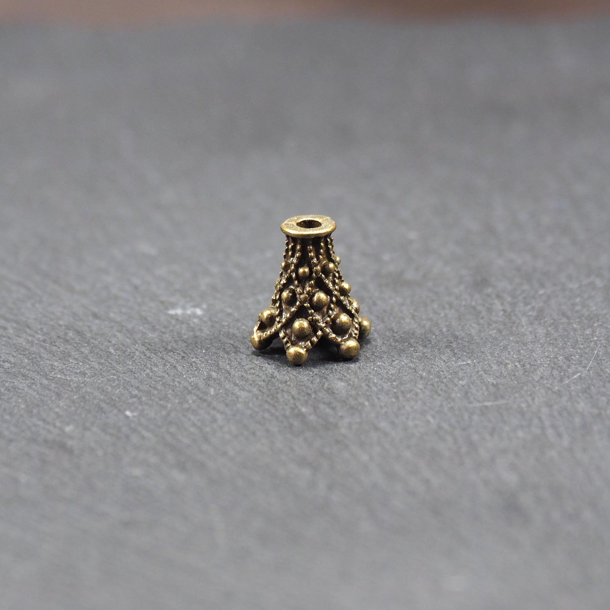 Konische bronzefarbene Perlenkappe aus Metall mit filigranem Ornamentreliéf, kugeligen Verzierungen und zentralem Fädelloch.