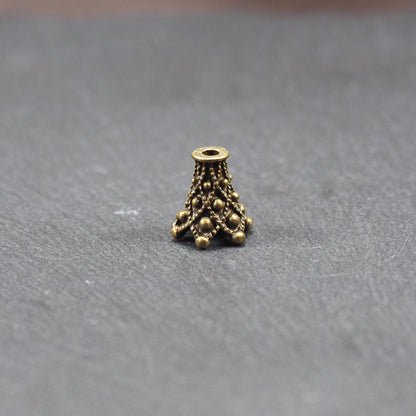 Konische bronzefarbene Perlenkappe aus Metall mit filigranem Ornamentreliéf, kugeligen Verzierungen und zentralem Fädelloch.