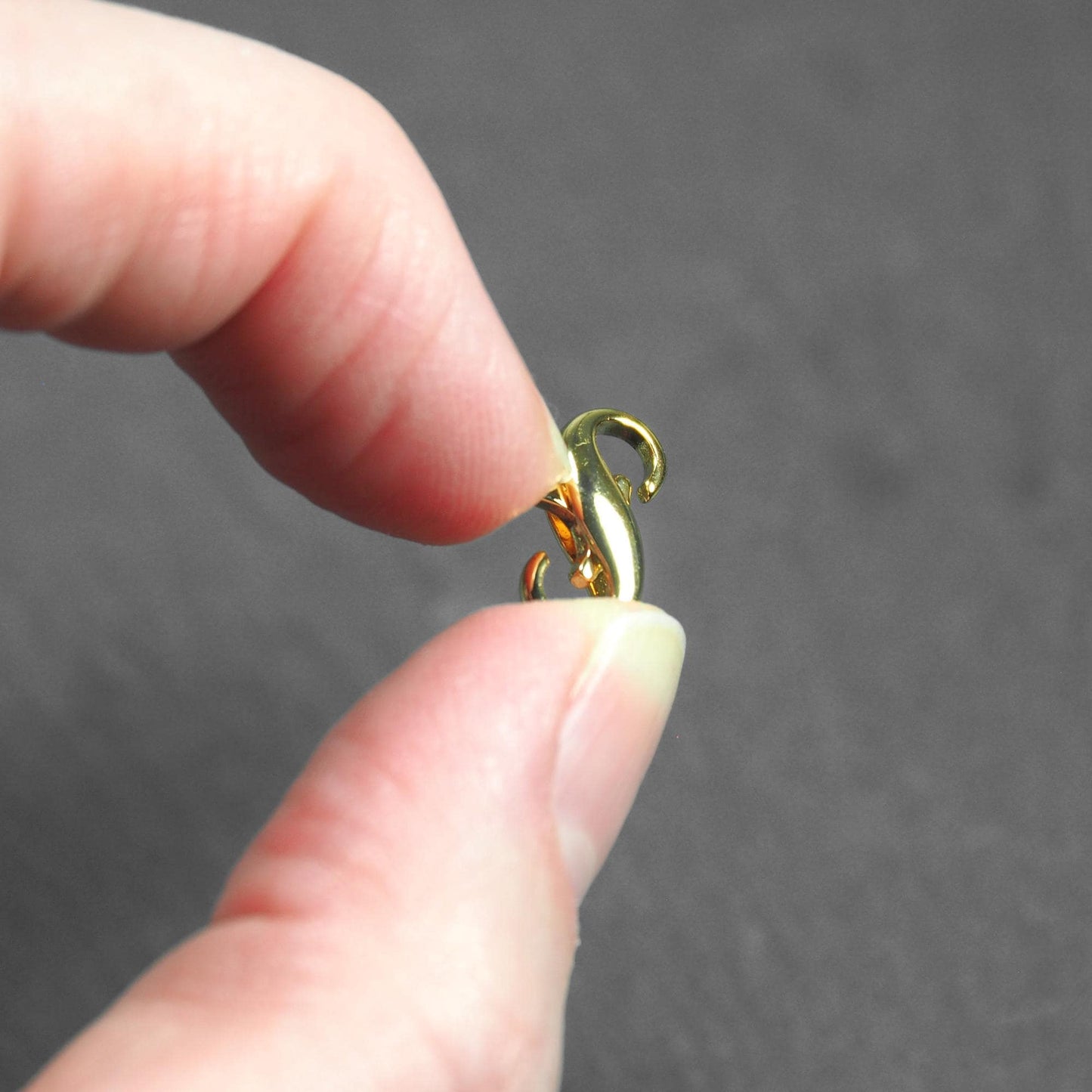 Goldfarbener Karabiner in liegender-Acht-Form mit zwei sichtbaren Ösen und glänzend glatter Oberfläche, zwischen Daumen und Zeigefinger gehalten.