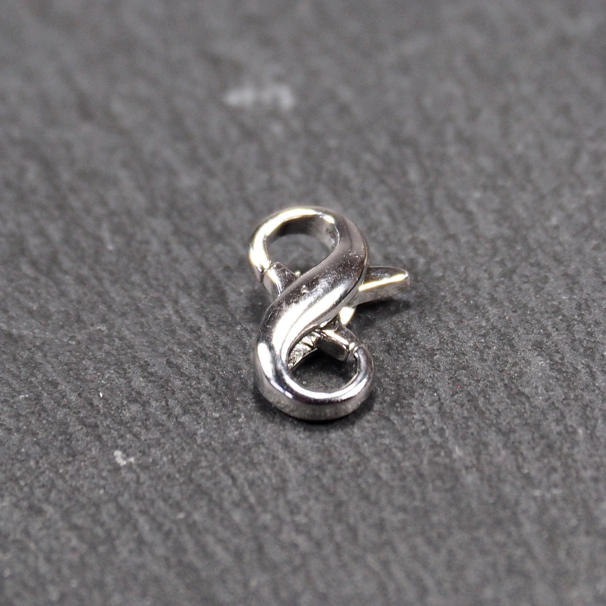 Ein doppelseitiger Karabiner im liegenden Acht-Design, silberfarben glänzend mit organisch geschwungener, durchbrochener Form und integrierter Öse.