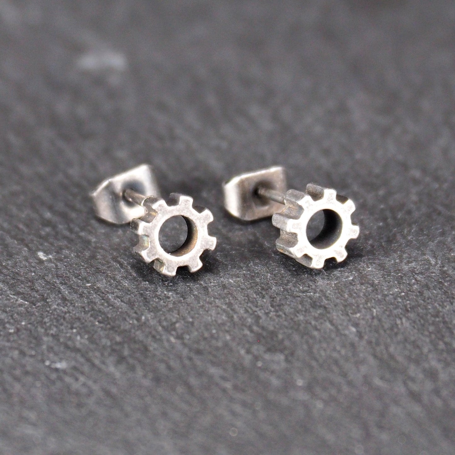 Ohrstecker in silberner, matter Metalloptik mit durchbrochenem Zahnrad-Motiv, zentraler Öffnung, kompaktem geometrischem Design und sichtbarem Steckstift.