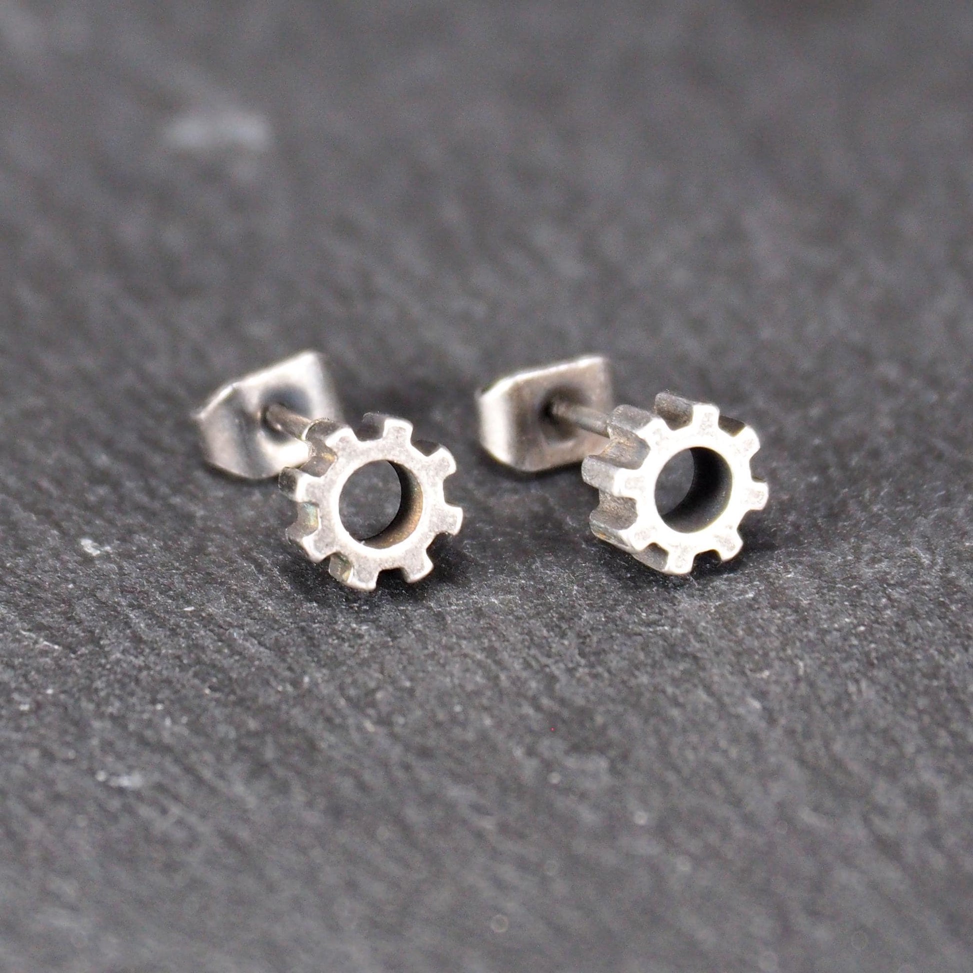 Ohrstecker in silberner, matter Metalloptik mit durchbrochenem Zahnrad-Motiv, zentraler Öffnung, kompaktem geometrischem Design und sichtbarem Steckstift.