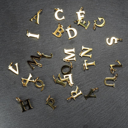 Mehrere goldfarbene glänzende Großbuchstaben-Anhänger in versaler Serifen-Schrift, klein und filigran, jeweils mit kleiner Öse.