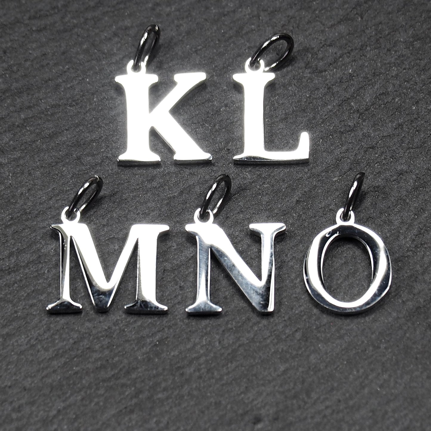 Buchstaben-Anhänger aus poliertem silberfarbenem Metall, serifenhafte Großbuchstaben K L M N O, jeweils mit kleiner Öse oben.