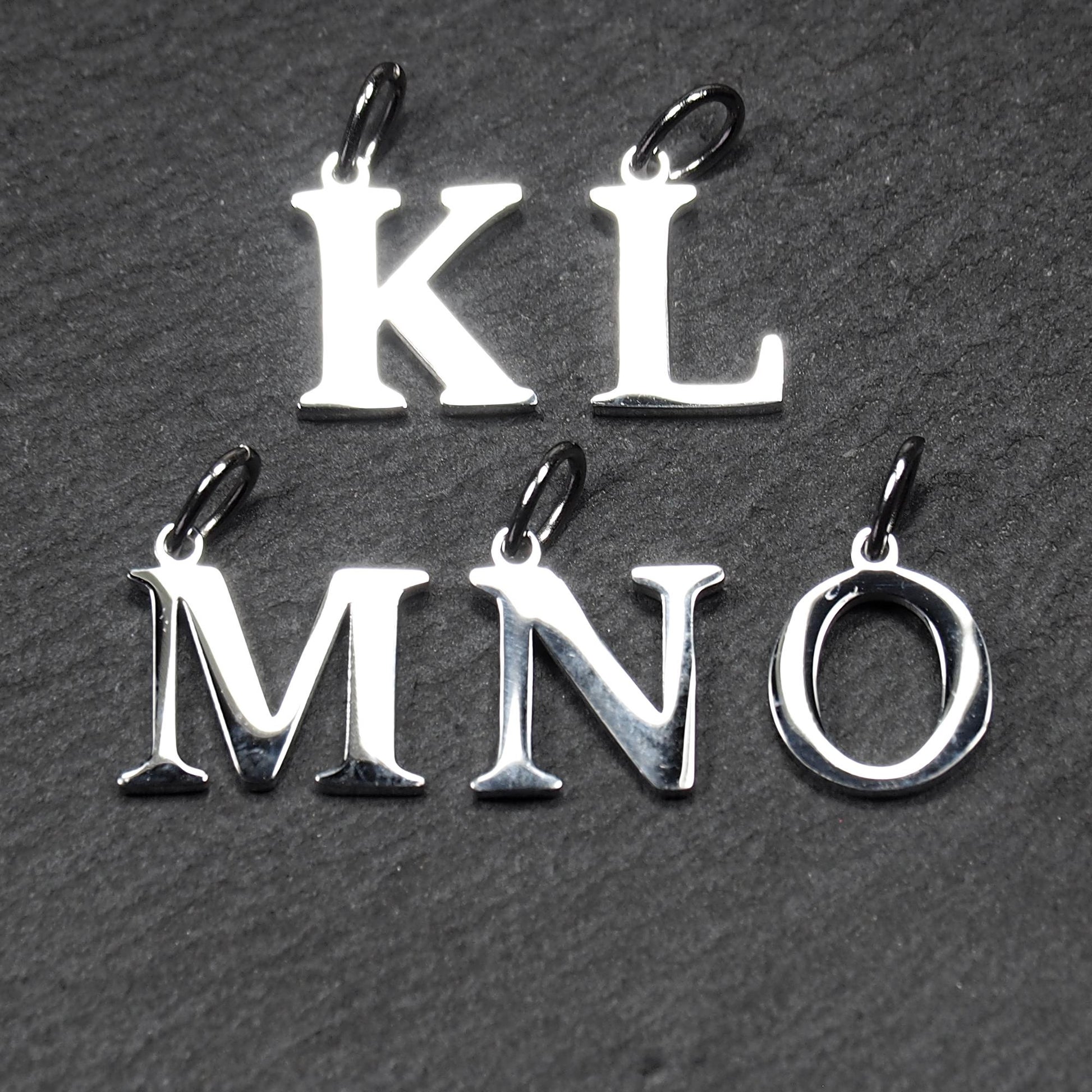Buchstaben-Anhänger aus poliertem silberfarbenem Metall, serifenhafte Großbuchstaben K L M N O, jeweils mit kleiner Öse oben.