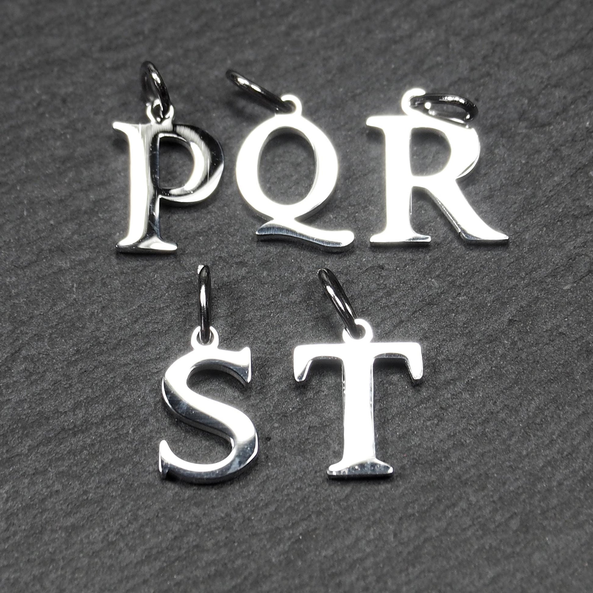 Fünf polierte, silberfarbene Buchstaben-Anhänger in Großbuchstaben (P, Q, R, S, T) mit Serifenschrift und obenliegender Öse auf dunklem strukturiertem Hintergrund.
