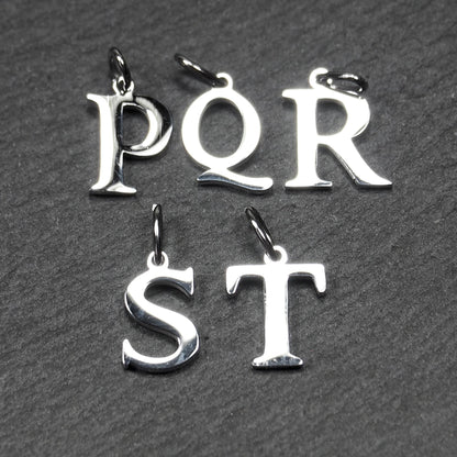 Fünf polierte, silberfarbene Buchstaben-Anhänger in Großbuchstaben (P, Q, R, S, T) mit Serifenschrift und obenliegender Öse auf dunklem strukturiertem Hintergrund.