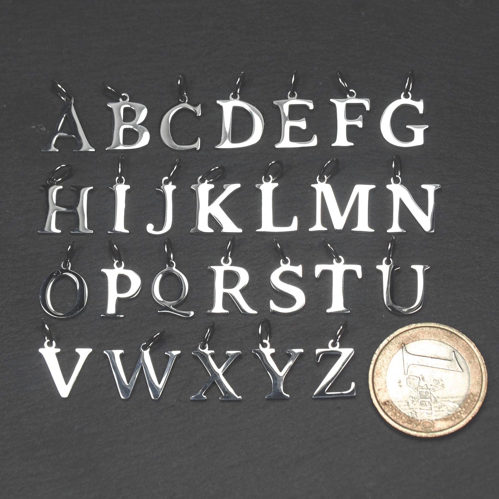 Großbuchstaben-Anhänger aus poliertem silberfarbenem Metall mit Serifen und kleiner Öse, angeordnet neben einer Ein-Euro-Münze.