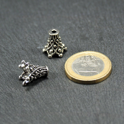 Zwei konische, antik-silberne, durchbrochene Perlenkappen mit filigranem Relief und kleinen Noppen, neben einer 1-Euro-Münze zur Größenanzeige.