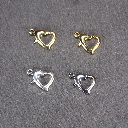 Vier herzförmige Karabiner-Verschlüsse mit sichtbarer Öse, zwei glänzend goldfarbene und zwei glänzend silberfarbene Exemplare auf dunklem Hintergrund.