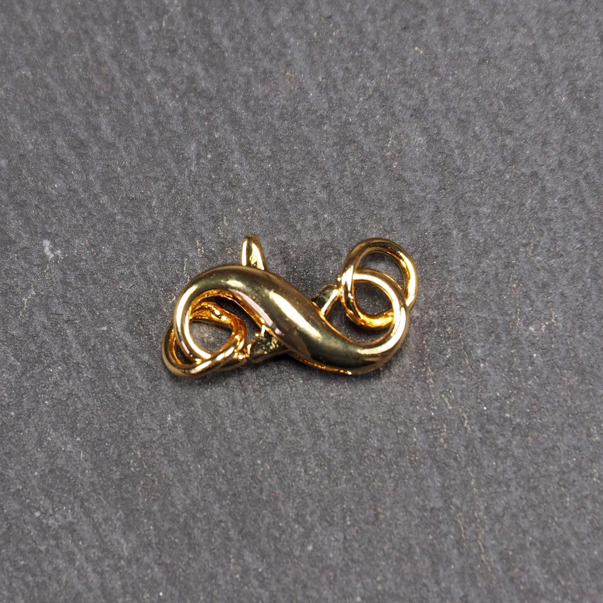 Karabiner in liegender Acht, goldfarbenes glänzendes Metall mit glatter Oberfläche, zwei gegenüberliegende Öffnungen und sichtbaren Ringösen.