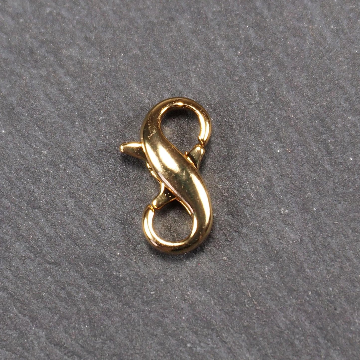 Karabiner in liegender Acht, goldfarbenes glänzendes Metall mit glatter Oberfläche, zwei gegenüberliegende Öffnungen und sichtbaren Ringösen.