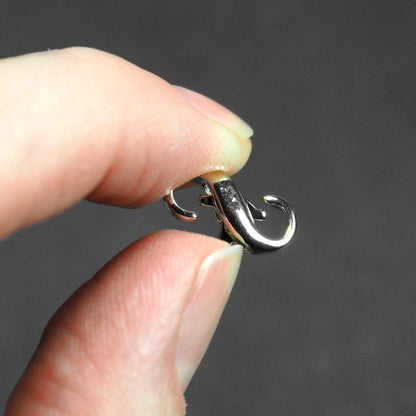 Doppelt öffnender Karabiner, silberglänzend mit organisch geschwungener liegender-Acht-Form und integrierter Öse, zwischen Daumen und Zeigefinger gehalten.