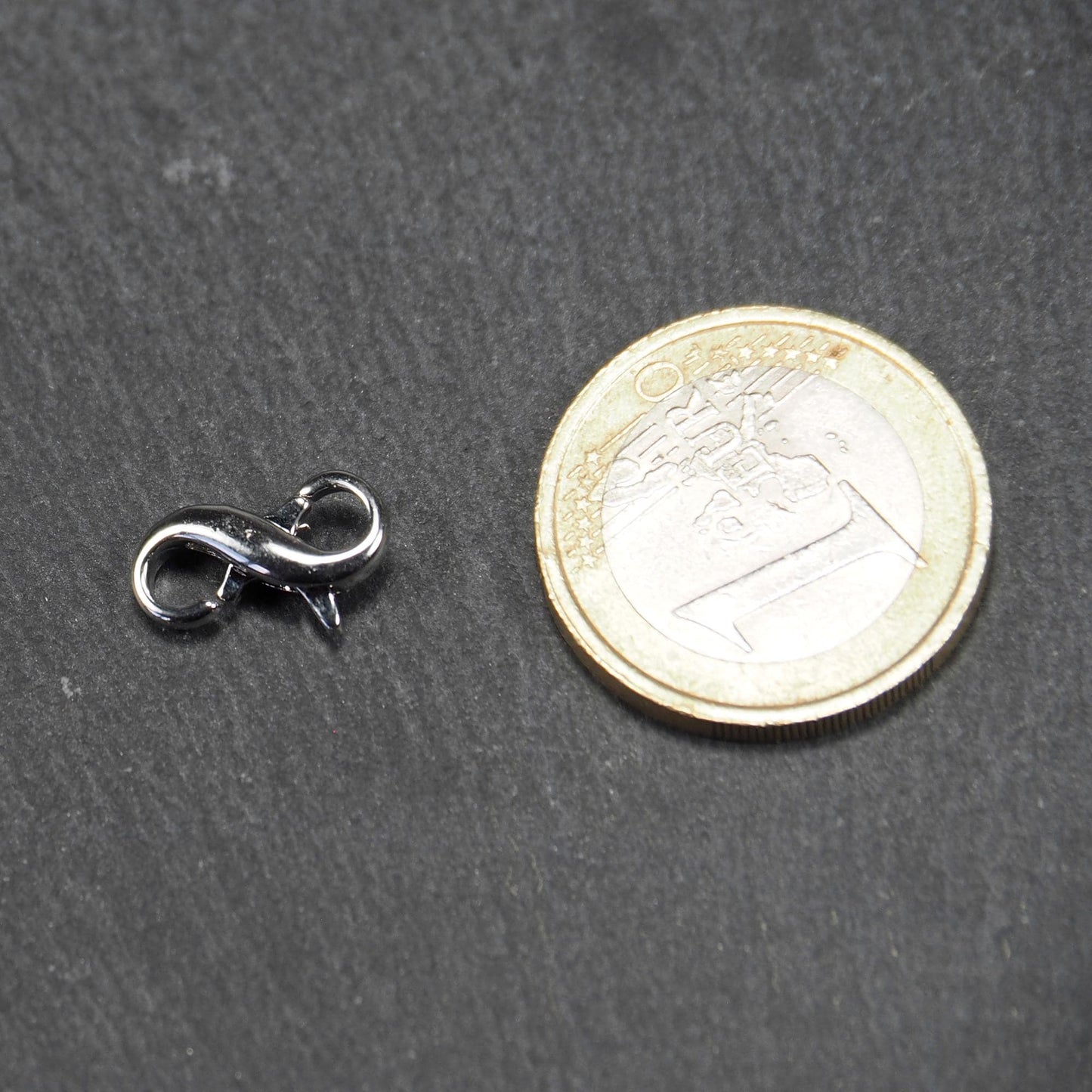 Ein doppelseitiger Karabiner im liegenden Acht-Design, silberfarben glänzend mit organisch geschwungener, durchbrochener Form und integrierter Öse.