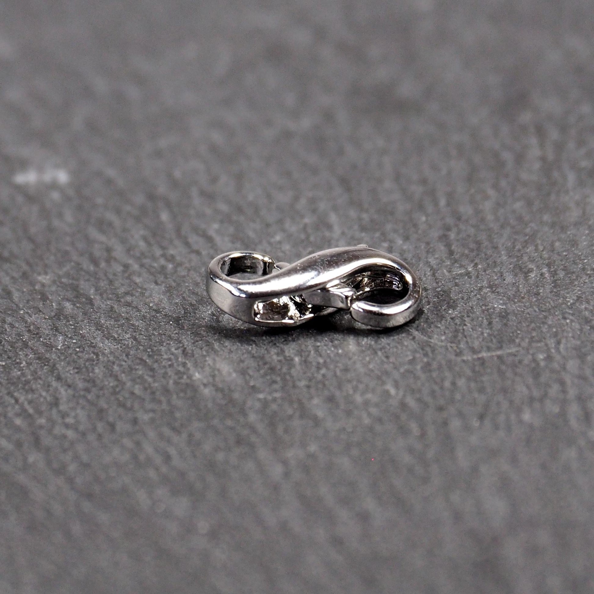 Ein doppelseitiger Karabiner im liegenden Acht-Design, silberfarben glänzend mit organisch geschwungener, durchbrochener Form und integrierter Öse.