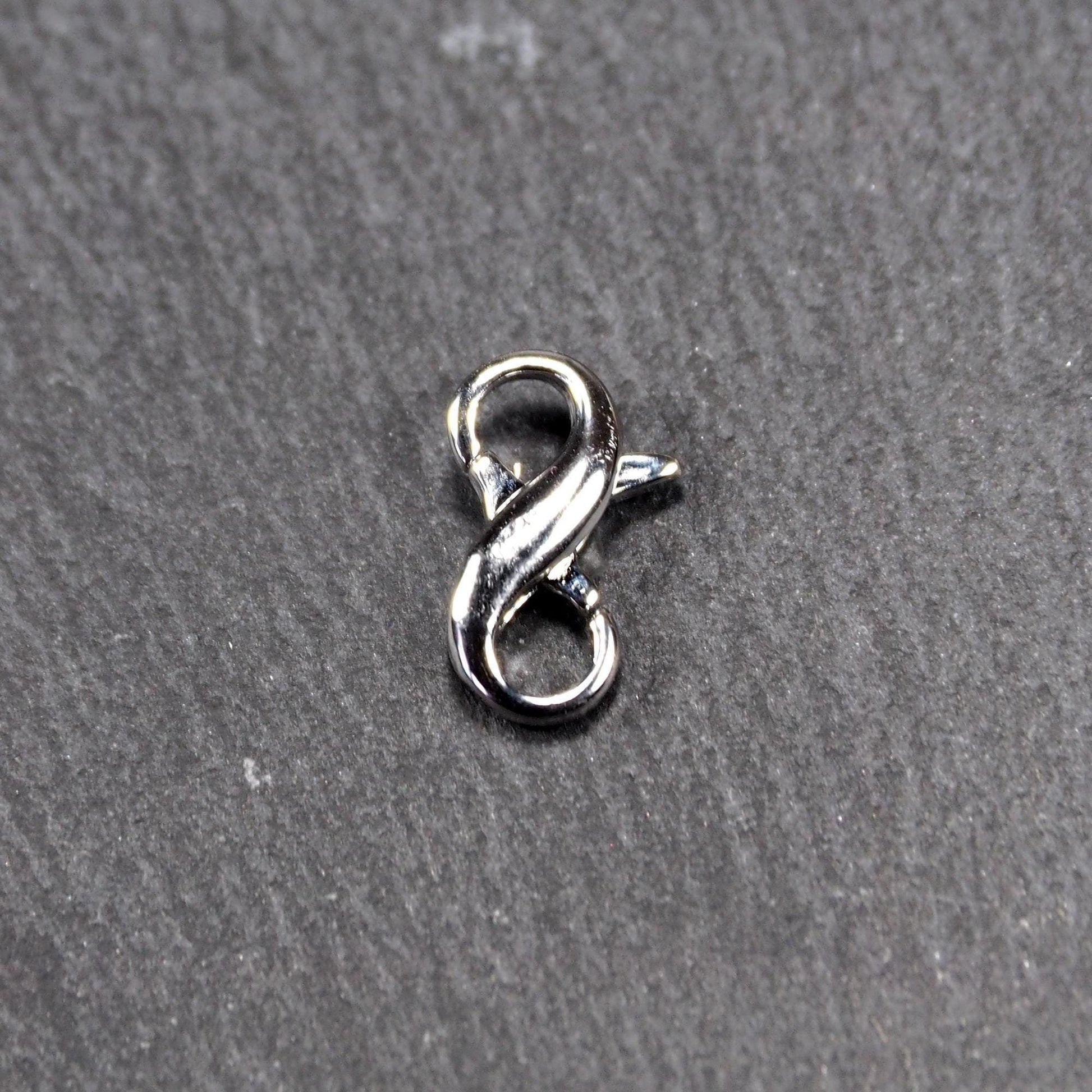 Ein doppelseitiger Karabiner im liegenden Acht-Design, silberfarben glänzend mit organisch geschwungener, durchbrochener Form und integrierter Öse.