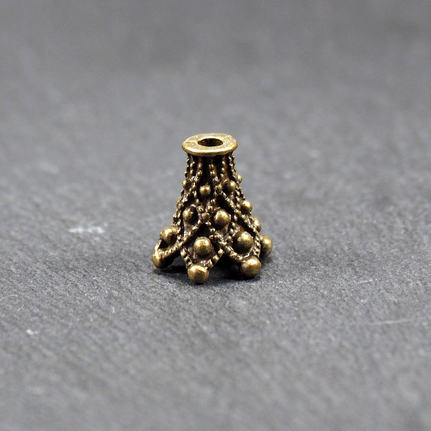 Konische bronzefarbene Perlenkappe aus Metall mit filigranem Ornamentreliéf, kugeligen Verzierungen und zentralem Fädelloch.