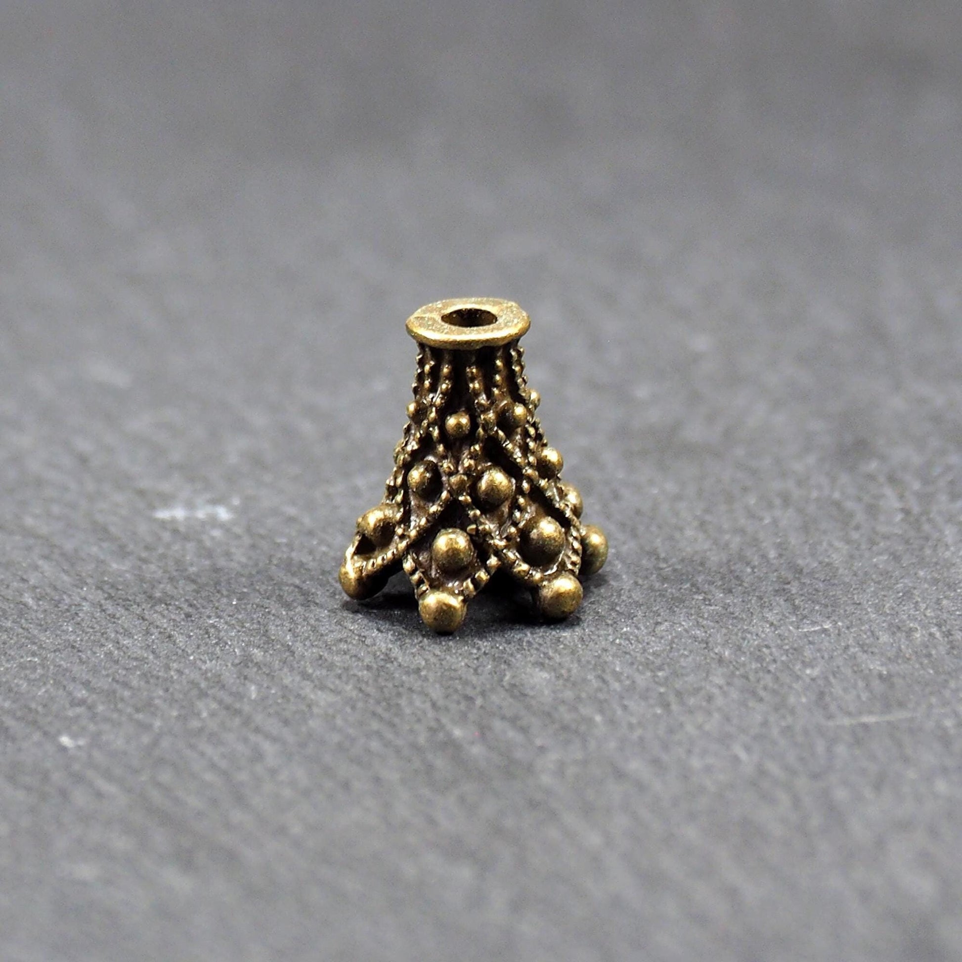Konische bronzefarbene Perlenkappe aus Metall mit filigranem Ornamentreliéf, kugeligen Verzierungen und zentralem Fädelloch.
