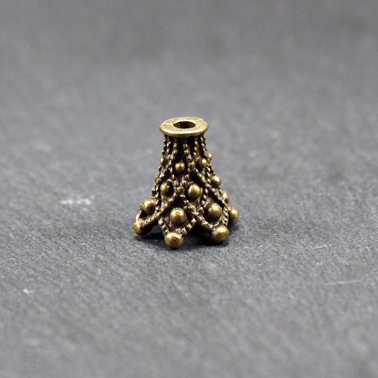 Konische bronzefarbene Perlenkappe aus Metall mit filigranem Ornamentreliéf, kugeligen Verzierungen und zentralem Fädelloch.