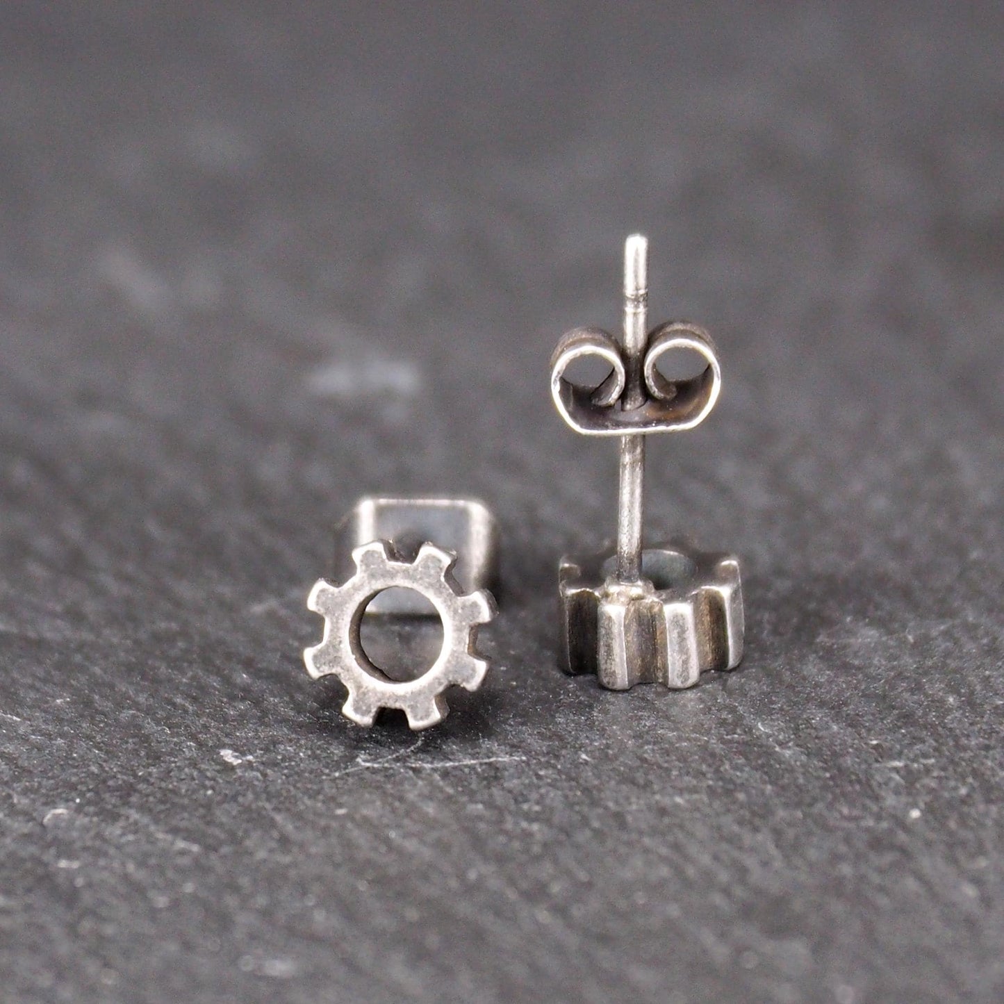 Ohrstecker in silberner, matter Metalloptik mit durchbrochenem Zahnrad-Motiv, zentraler Öffnung, kompaktem geometrischem Design und sichtbarem Steckstift.