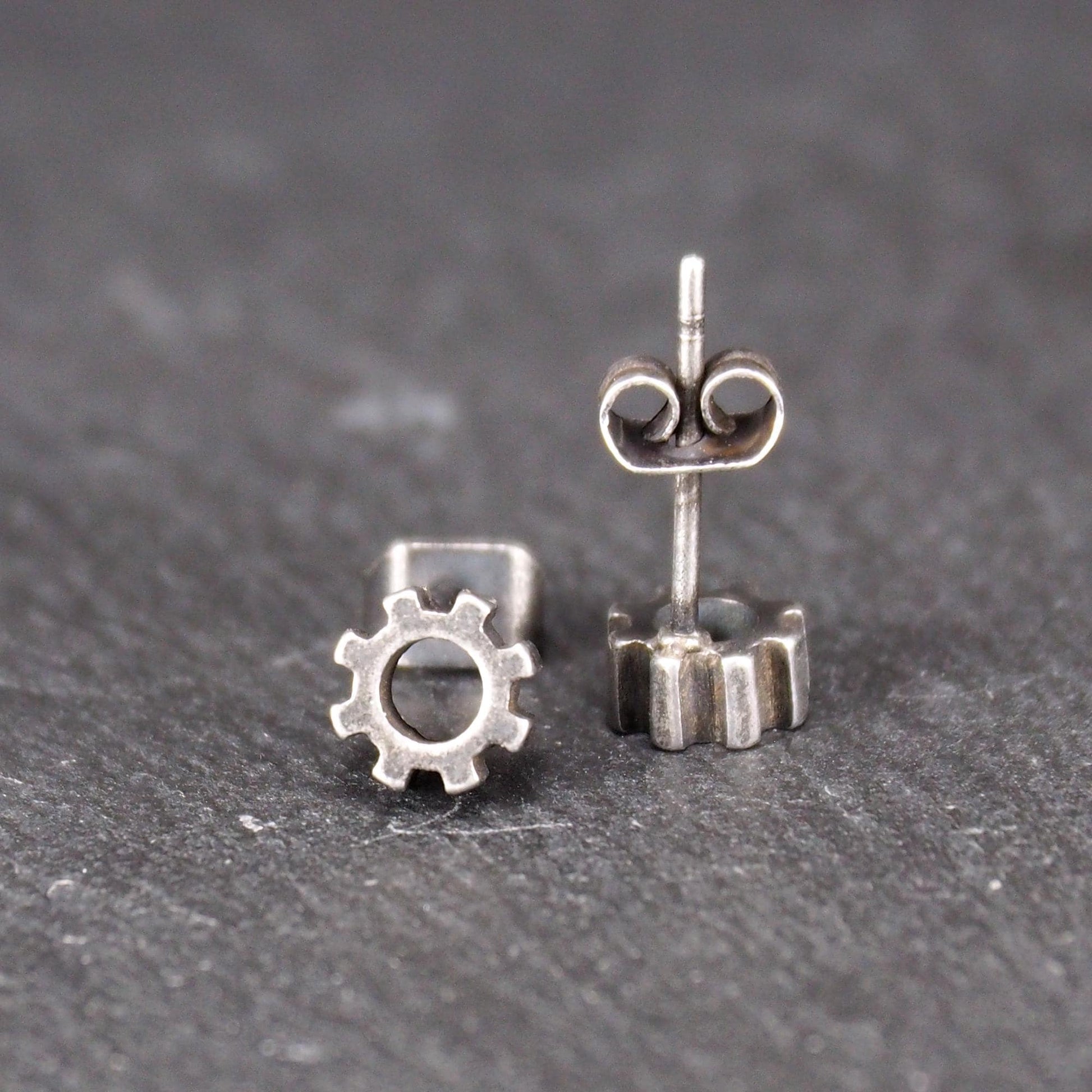 Ohrstecker in silberner, matter Metalloptik mit durchbrochenem Zahnrad-Motiv, zentraler Öffnung, kompaktem geometrischem Design und sichtbarem Steckstift.