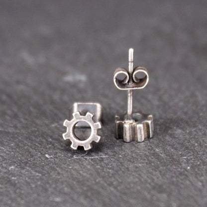 Ohrstecker in silberner, matter Metalloptik mit durchbrochenem Zahnrad-Motiv, zentraler Öffnung, kompaktem geometrischem Design und sichtbarem Steckstift.