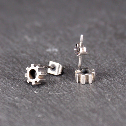 Ohrstecker in silberner, matter Metalloptik mit durchbrochenem Zahnrad-Motiv, zentraler Öffnung, kompaktem geometrischem Design und sichtbarem Steckstift.