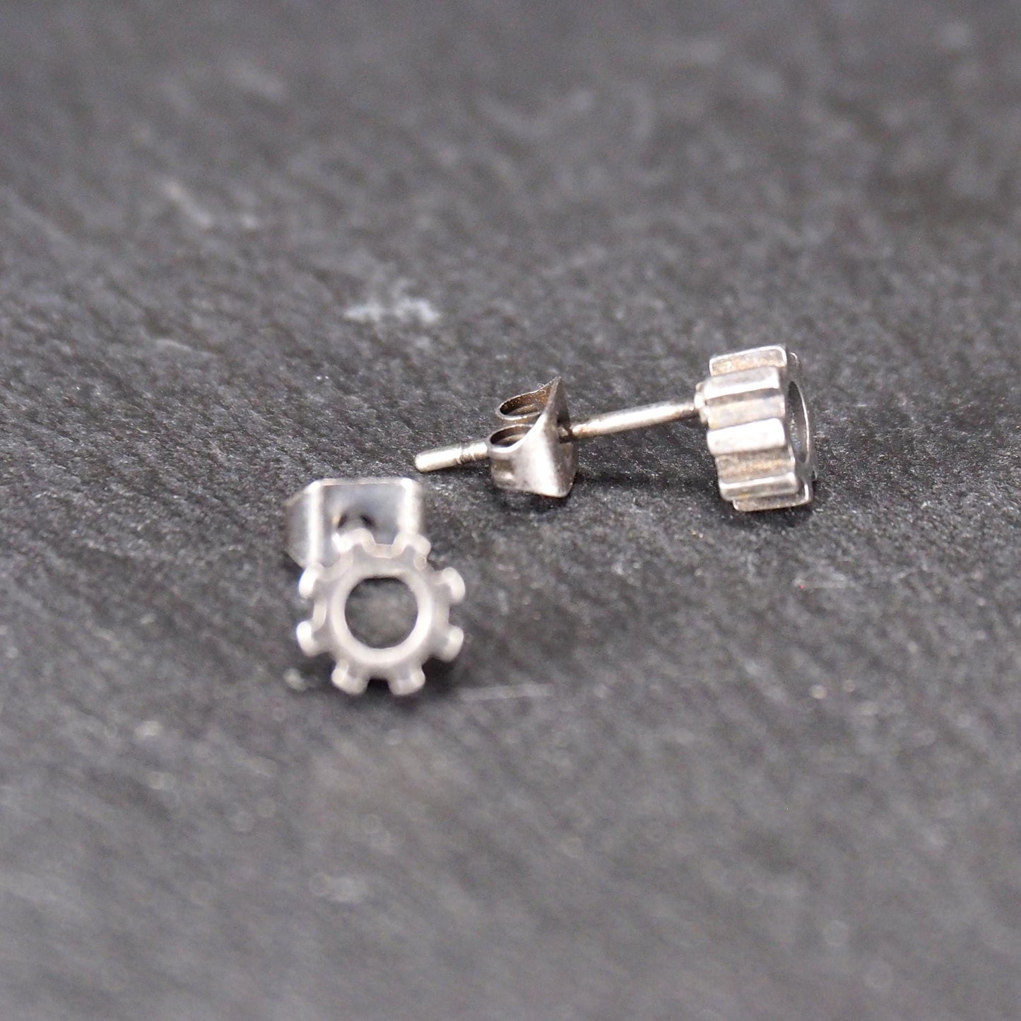 Ohrstecker in silberner, matter Metalloptik mit durchbrochenem Zahnrad-Motiv, zentraler Öffnung, kompaktem geometrischem Design und sichtbarem Steckstift.