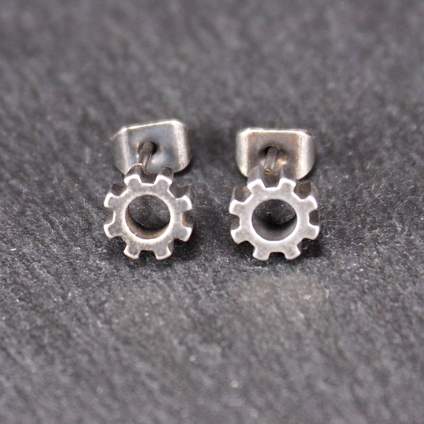 Ohrstecker in silberner, matter Metalloptik mit durchbrochenem Zahnrad-Motiv, zentraler Öffnung, kompaktem geometrischem Design und sichtbarem Steckstift.