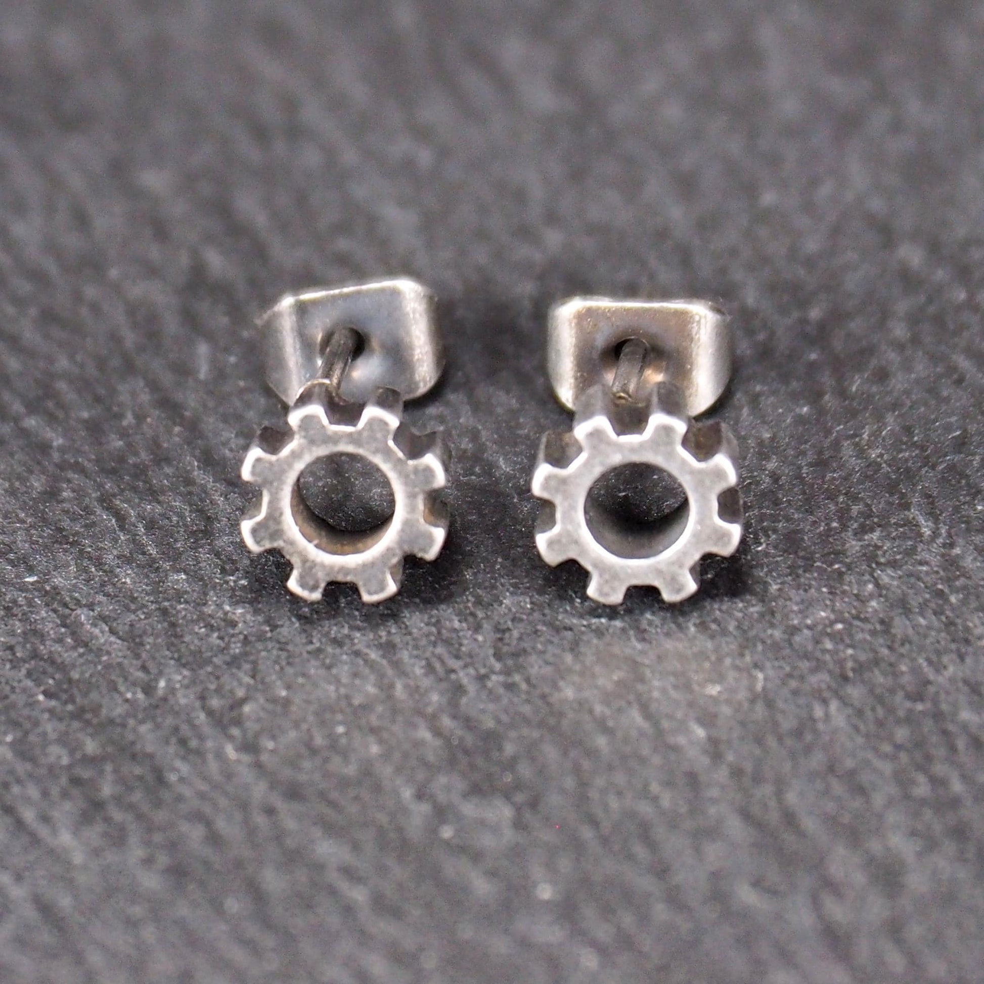 Ohrstecker in silberner, matter Metalloptik mit durchbrochenem Zahnrad-Motiv, zentraler Öffnung, kompaktem geometrischem Design und sichtbarem Steckstift.