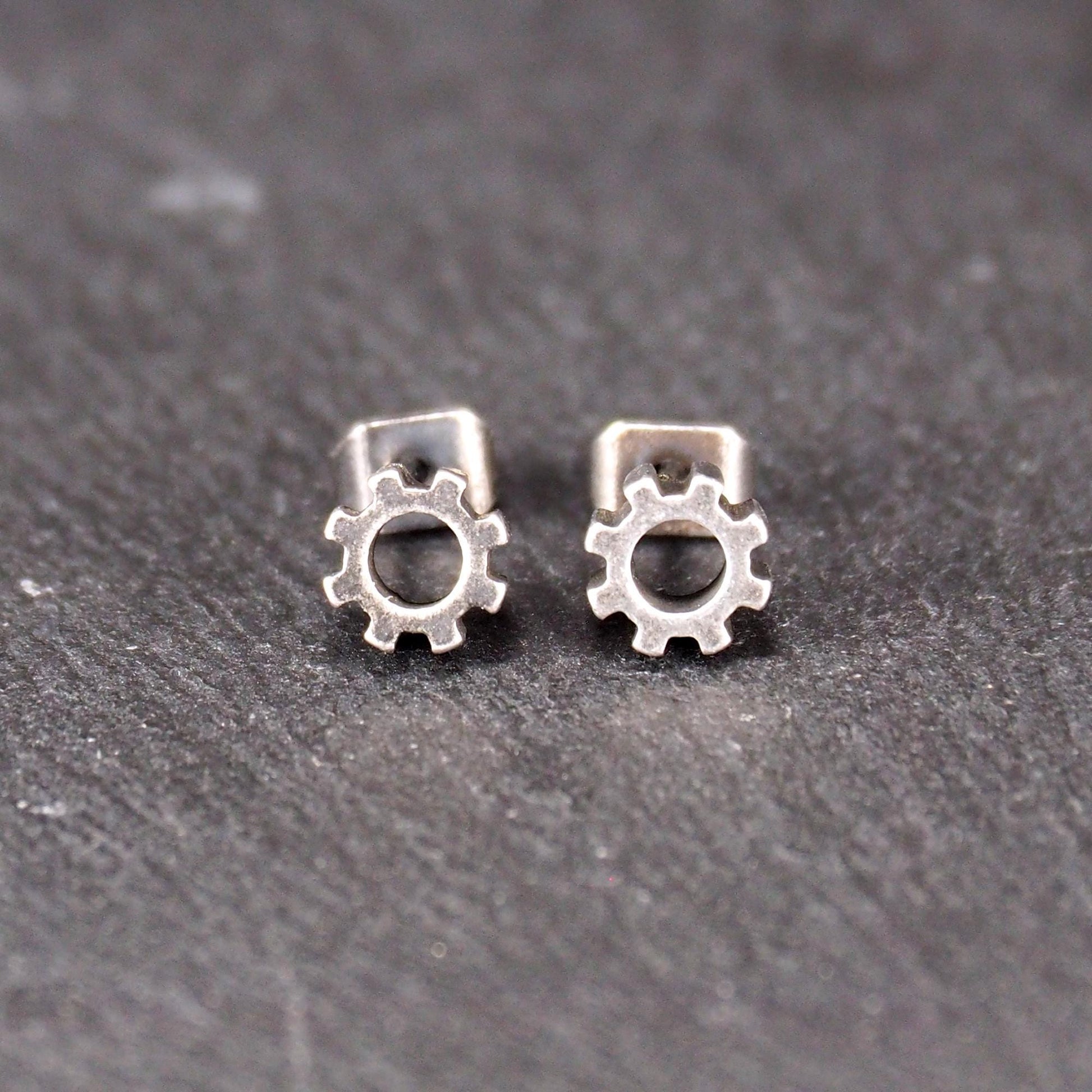 Ohrstecker in silberner, matter Metalloptik mit durchbrochenem Zahnrad-Motiv, zentraler Öffnung, kompaktem geometrischem Design und sichtbarem Steckstift.