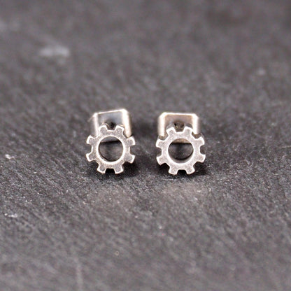 Ohrstecker in silberner, matter Metalloptik mit durchbrochenem Zahnrad-Motiv, zentraler Öffnung, kompaktem geometrischem Design und sichtbarem Steckstift.