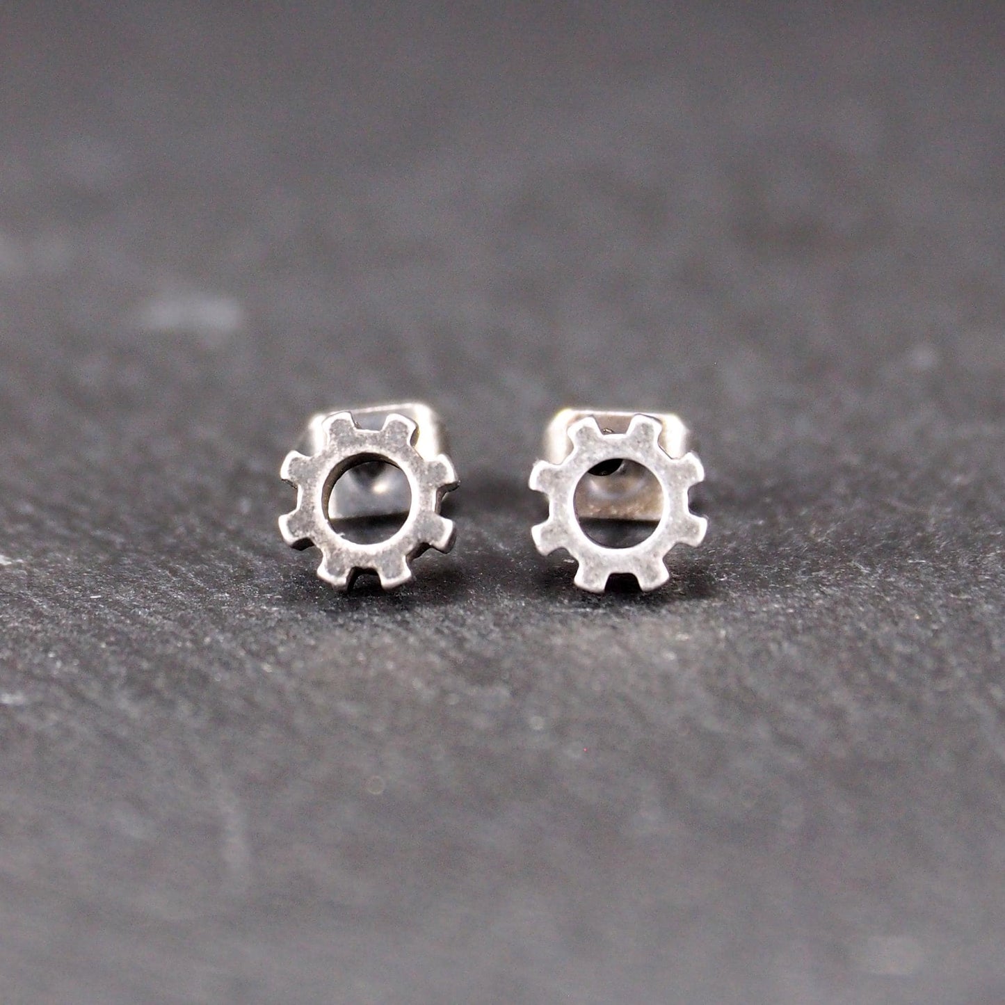 Ohrstecker in silberner, matter Metalloptik mit durchbrochenem Zahnrad-Motiv, zentraler Öffnung, kompaktem geometrischem Design und sichtbarem Steckstift.