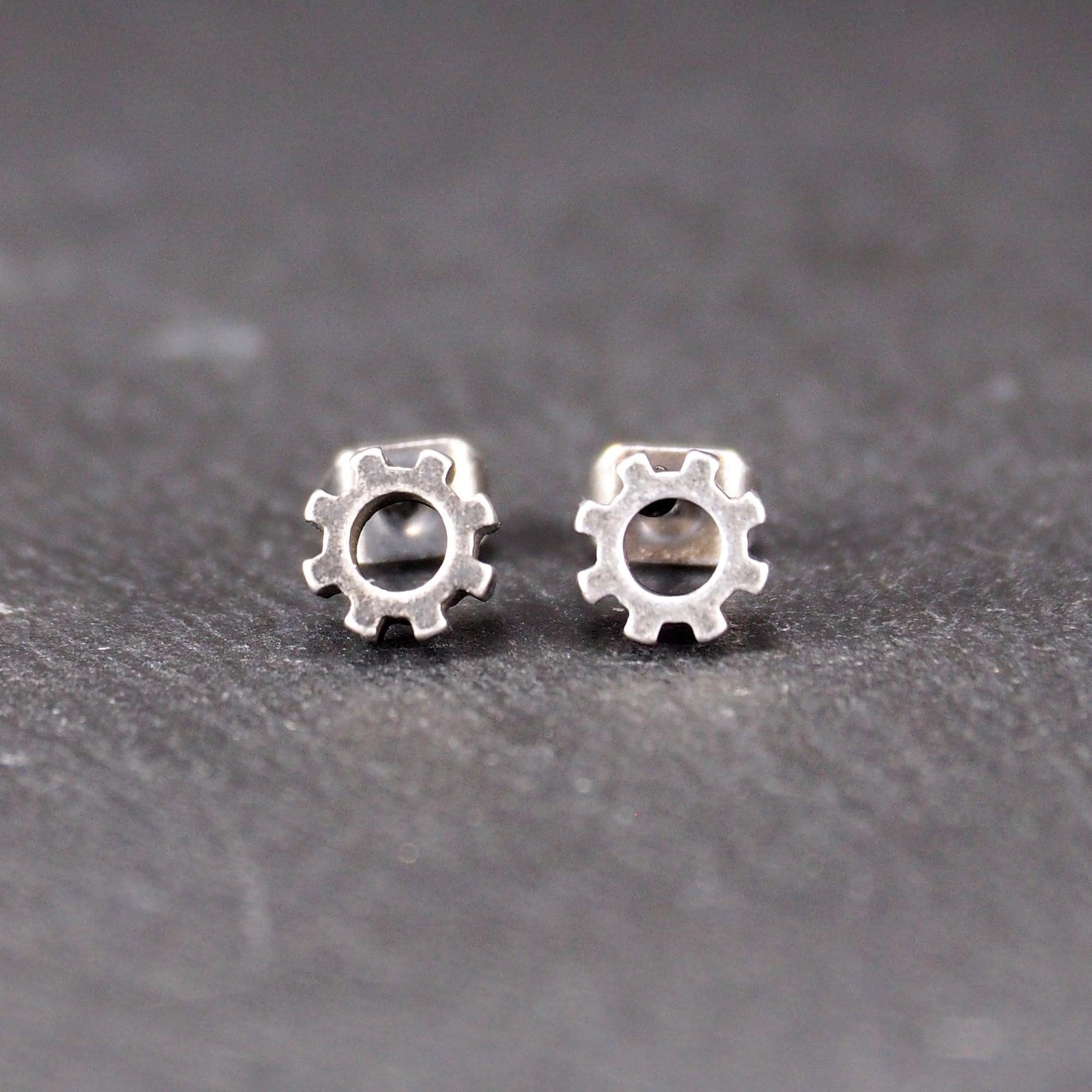 Ohrstecker in silberner, matter Metalloptik mit durchbrochenem Zahnrad-Motiv, zentraler Öffnung, kompaktem geometrischem Design und sichtbarem Steckstift.