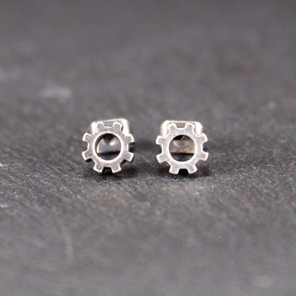 Ohrstecker in silberner, matter Metalloptik mit durchbrochenem Zahnrad-Motiv, zentraler Öffnung, kompaktem geometrischem Design und sichtbarem Steckstift.