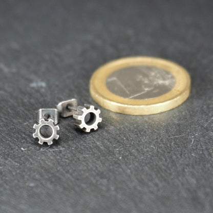 Ohrstecker, matte silberne Zahnräder mit rundem Mittelloch, Steg mit Steckverschluss; neben einer Euromünze zum Größenvergleich.