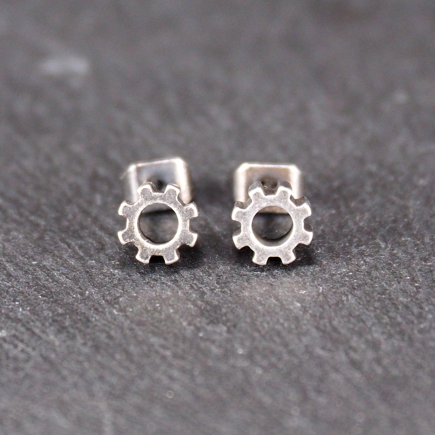 Ohrstecker in silberner, matter Metalloptik mit durchbrochenem Zahnrad-Motiv, zentraler Öffnung, kompaktem geometrischem Design und sichtbarem Steckstift.