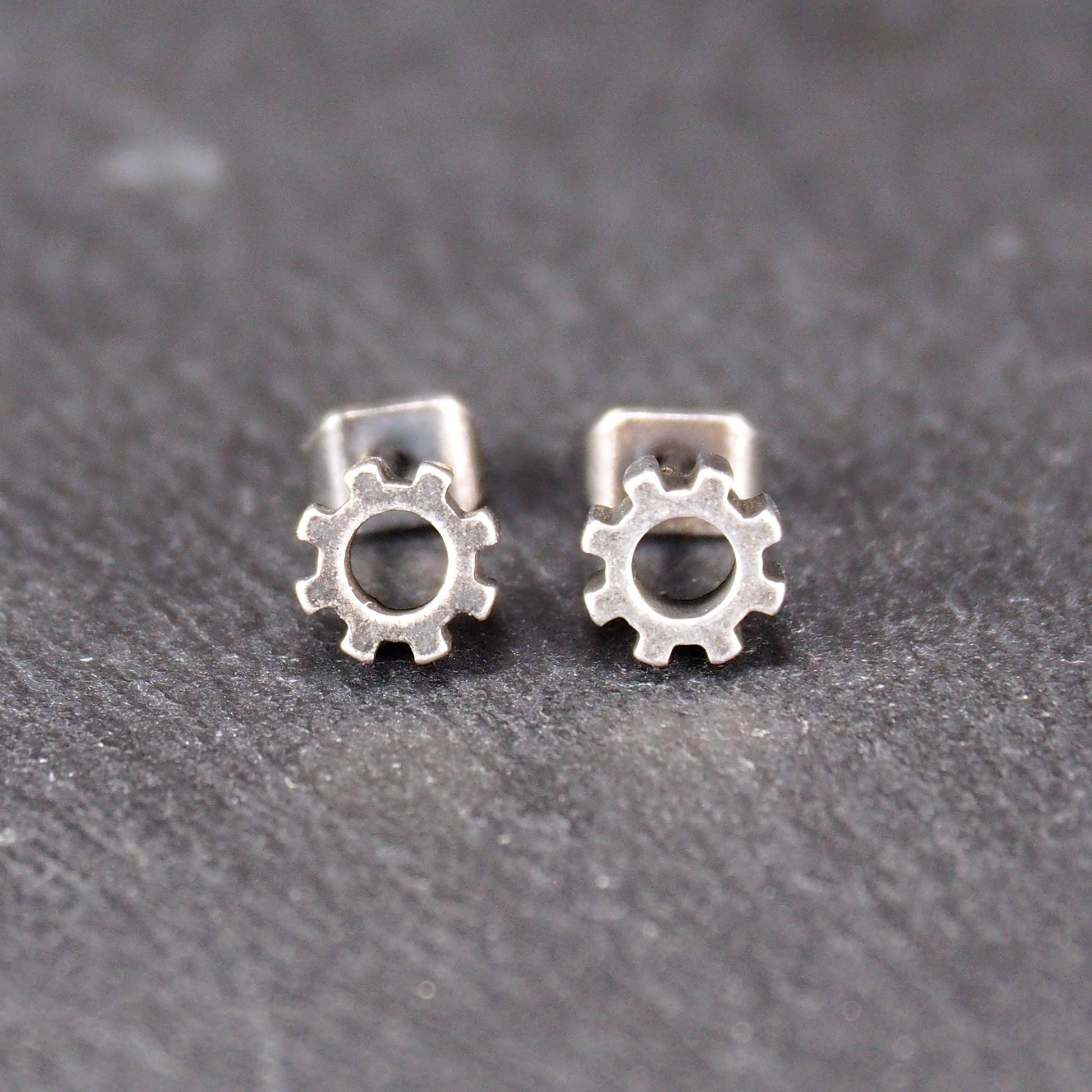 Ohrstecker in silberner, matter Metalloptik mit durchbrochenem Zahnrad-Motiv, zentraler Öffnung, kompaktem geometrischem Design und sichtbarem Steckstift.