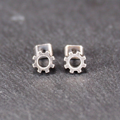 Ohrstecker in silberner, matter Metalloptik mit durchbrochenem Zahnrad-Motiv, zentraler Öffnung, kompaktem geometrischem Design und sichtbarem Steckstift.