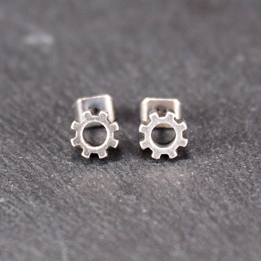 Ohrstecker in silberner, matter Metalloptik mit durchbrochenem Zahnrad-Motiv, zentraler Öffnung, kompaktem geometrischem Design und sichtbarem Steckstift.