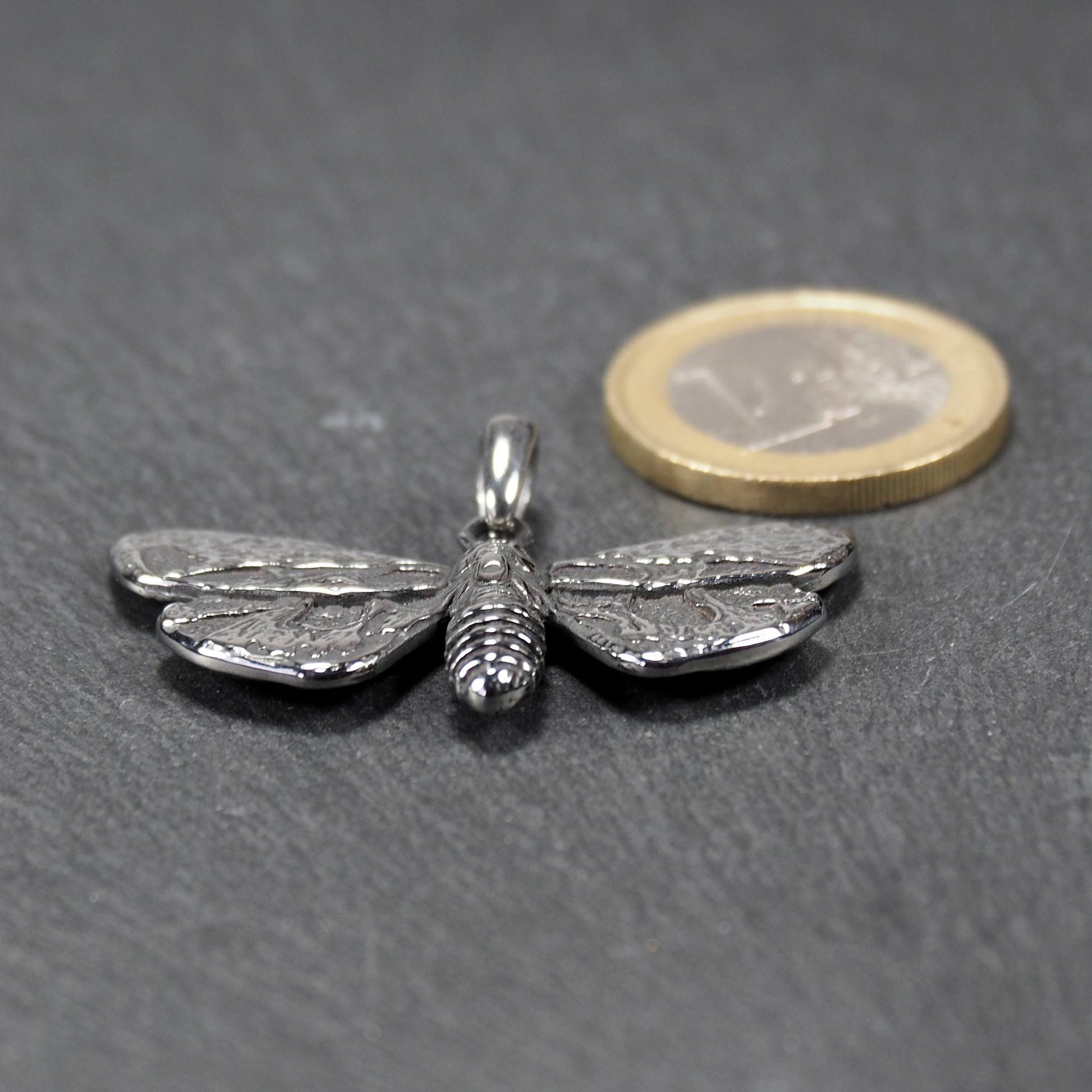 Anhänger in Mottenform, silberfarbene glänzende Metalloberfläche mit reliefartigen, strukturierten Flügeln, segmentiertem Körper und integrierter Öse. Mit 1 Euro Münze zum Größenvergleich.